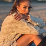 Jennifer Lopez sitting on a beach during sunset wearing a cream knit sweater and black boots