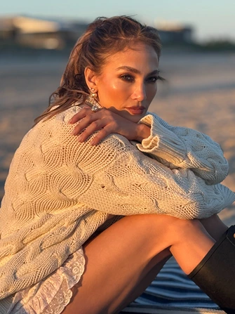 Jennifer Lopez sitting on a beach during sunset wearing a cream knit sweater and black boots
