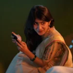 Woman wearing a traditional saree during Pongal celebrations, smiling while holding a mobile phone in warm festive lighting.