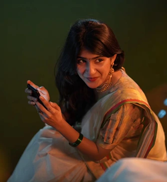 Woman wearing a traditional saree during Pongal celebrations, smiling while holding a mobile phone in warm festive lighting.